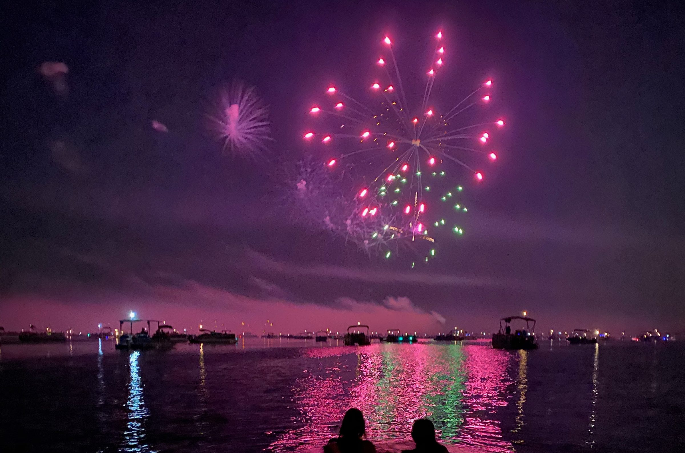 Two swimmers watching fireworks over Lake Lotawana, their reflections glowing pink and green in the water